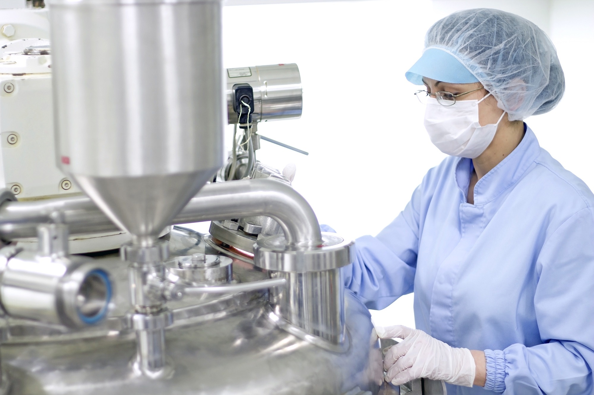 A technician in a lab coat, hairnet, and mask operates stainless steel machinery, illustrating a cleanroom environment for precision work.