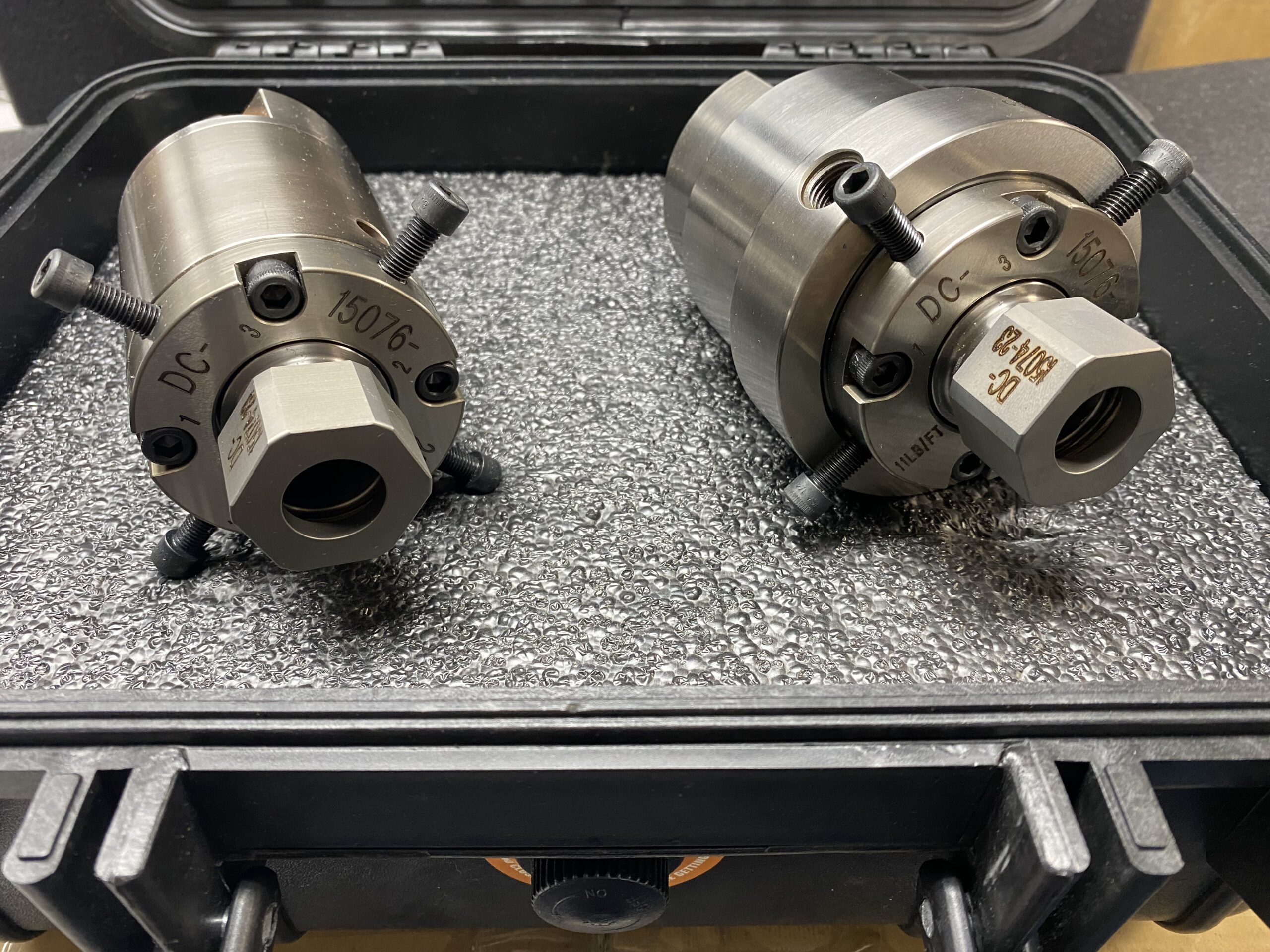 Two metal cylindrical tools with bolts and engraved markings are placed inside a textured foam-lined black case, suggesting industrial usage.