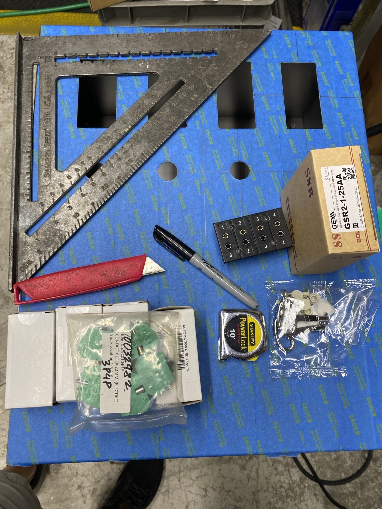 Tools and supplies laid out on a blue covered work surface, including a square, utility knife, tape measure, and various electrical components for a project.