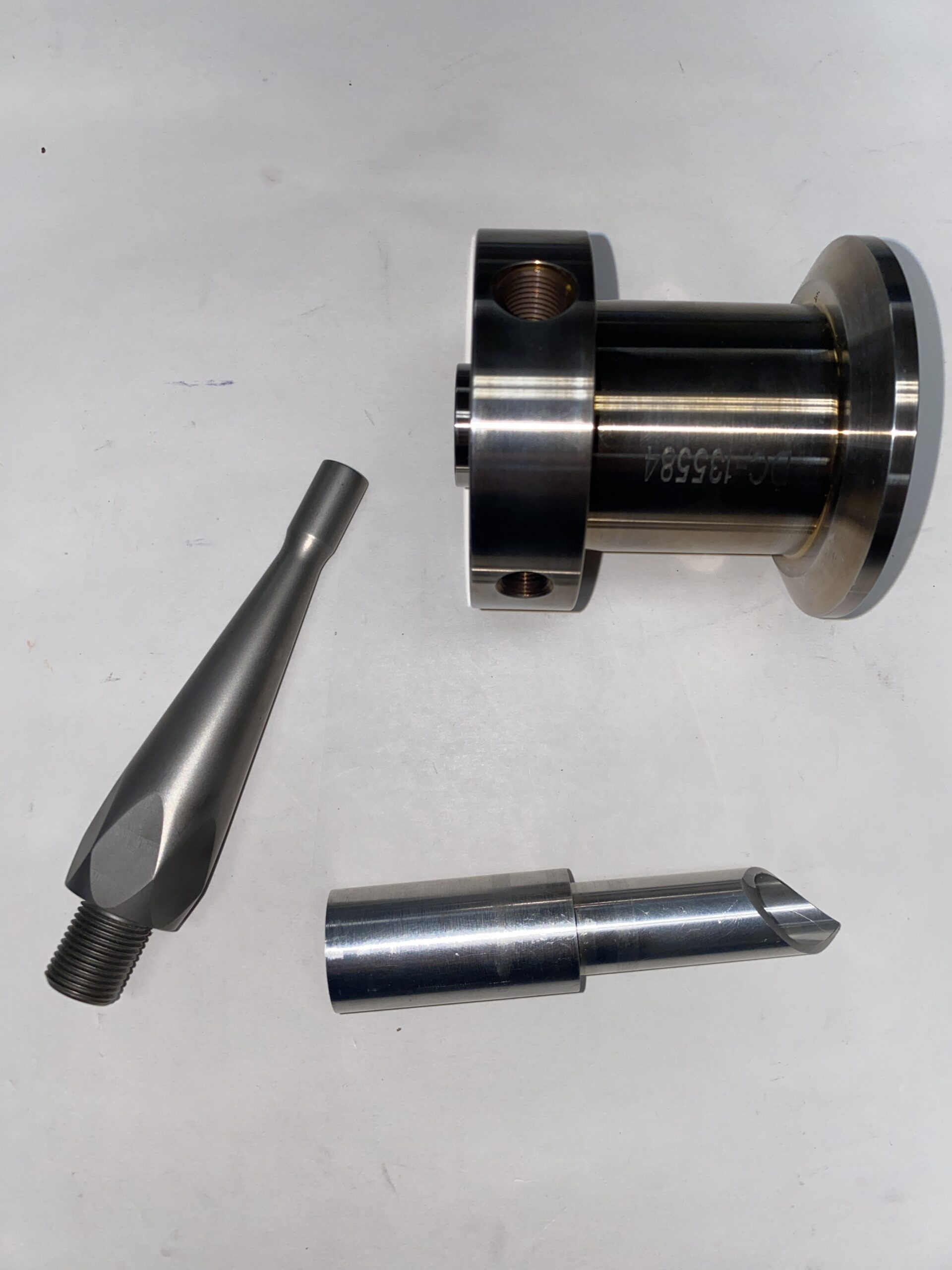 Three precision metal parts are displayed: a cylindrical adaptor with a flange, a slender nozzle, and a tapered tool, showcasing machining components.
